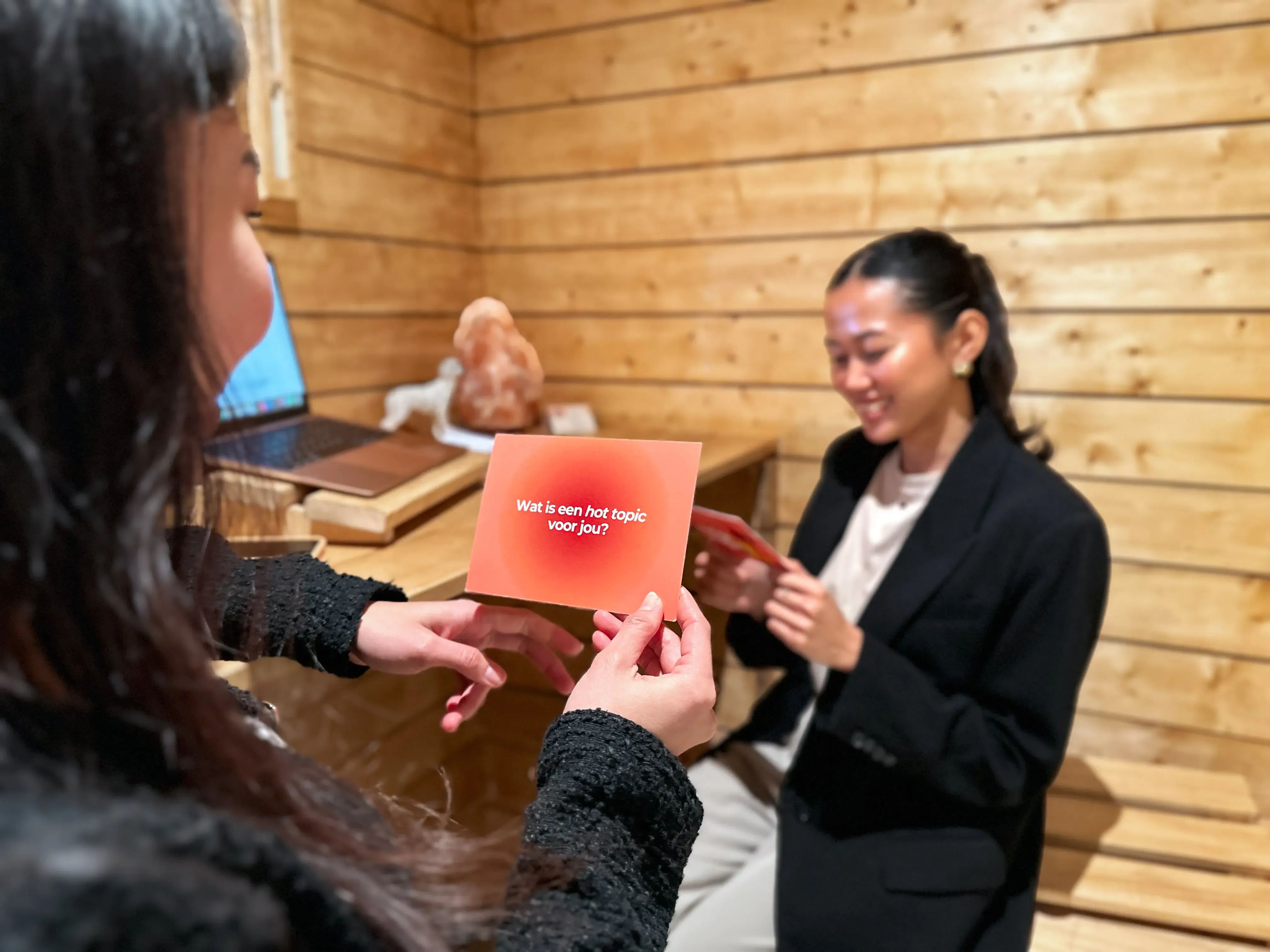 Vrouw houdt een oranje kaartje met een vraag omhoog naar een glimlachende vriendin.
