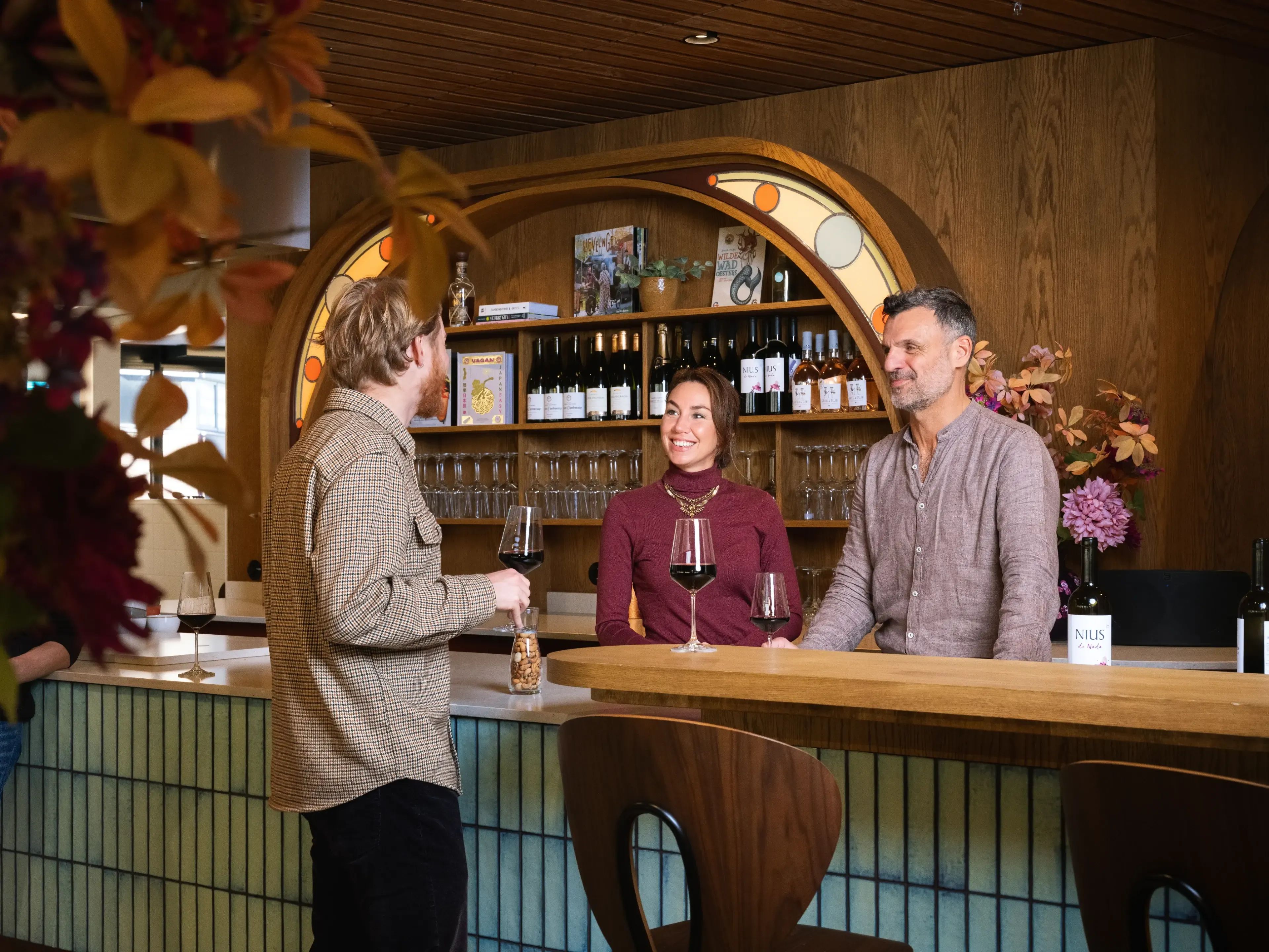 Drie mensen in een moderne wijnbar, twee achter de toonbank, één aan het wijn proeven.