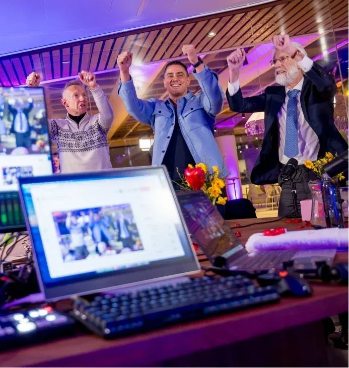 Drie mannen vieren feest met opgeheven armen achter een laptop waarop een groepsfoto te zien is.