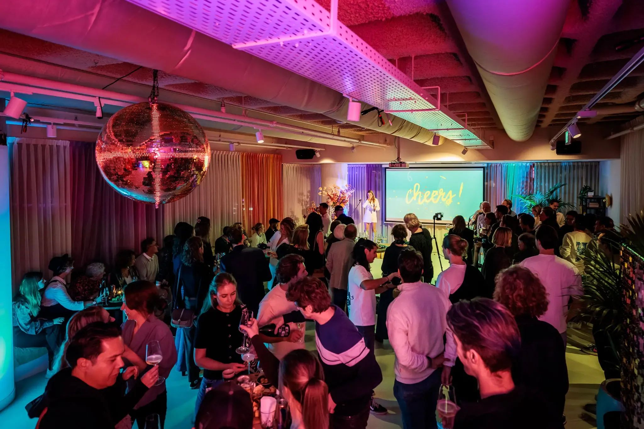 Binnenevenement met een discobal, kleurrijke verlichting en veel mensen die met elkaar socializen. 