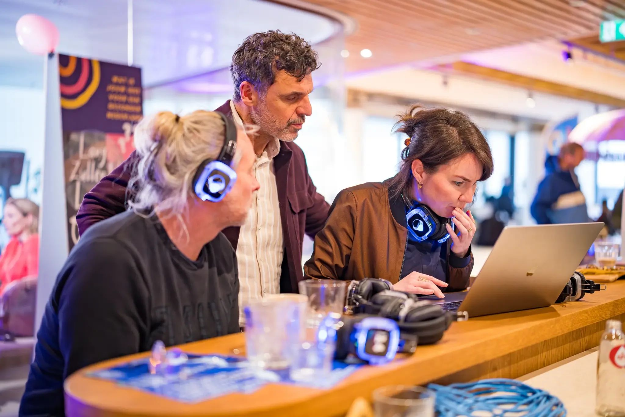 Drie mensen met lichtgevende koptelefoons kijken aandachtig naar een laptopscherm aan een balie.