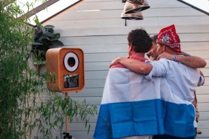 Drie mannen, waarvan één met een Luxemburgse vlag, poseren samen bij een oranje fotohokje in de open lucht.