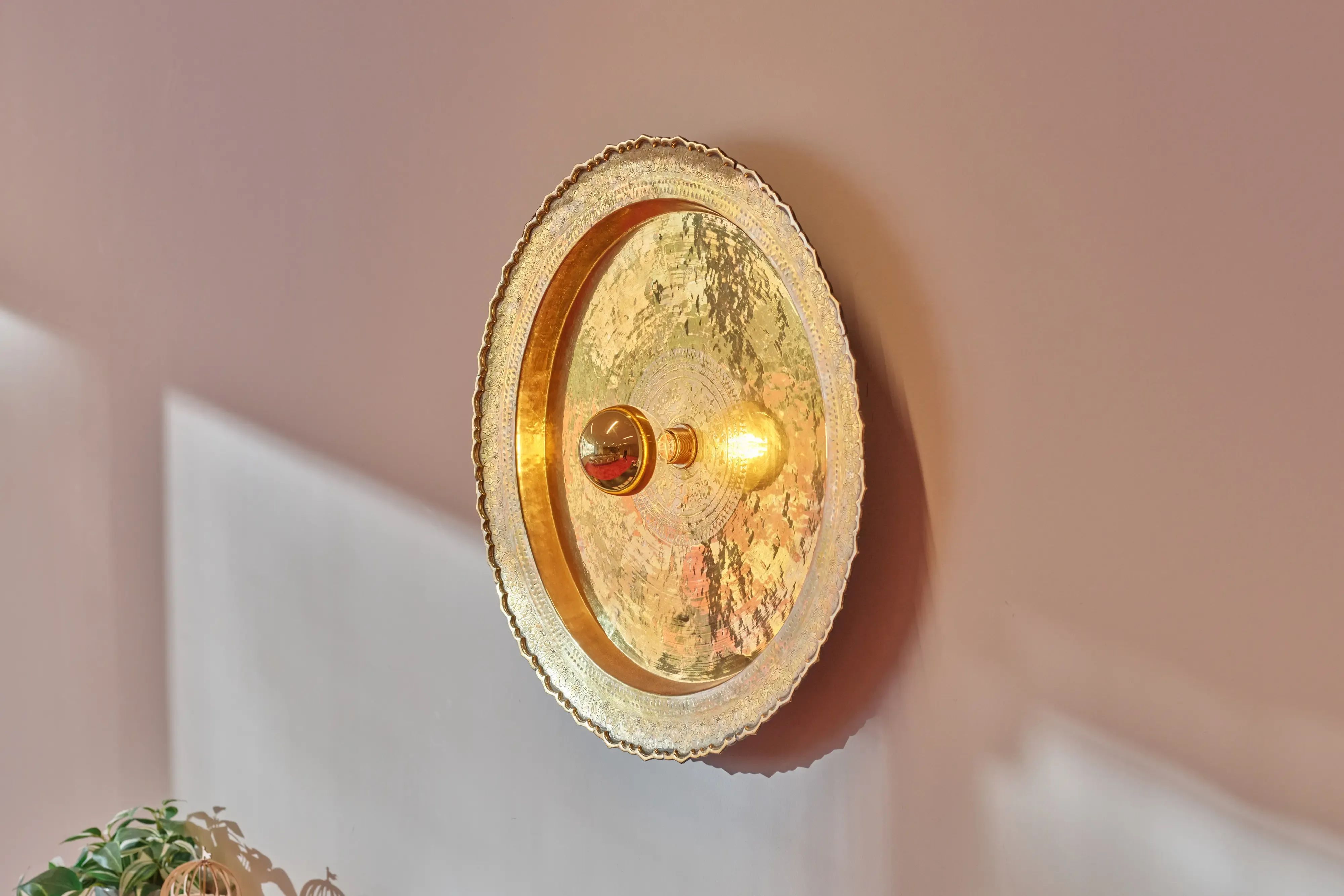 Een ronde, gehamerde gouden wandlamp met een gloeiende lamp gemonteerd op een lichtroze muur.