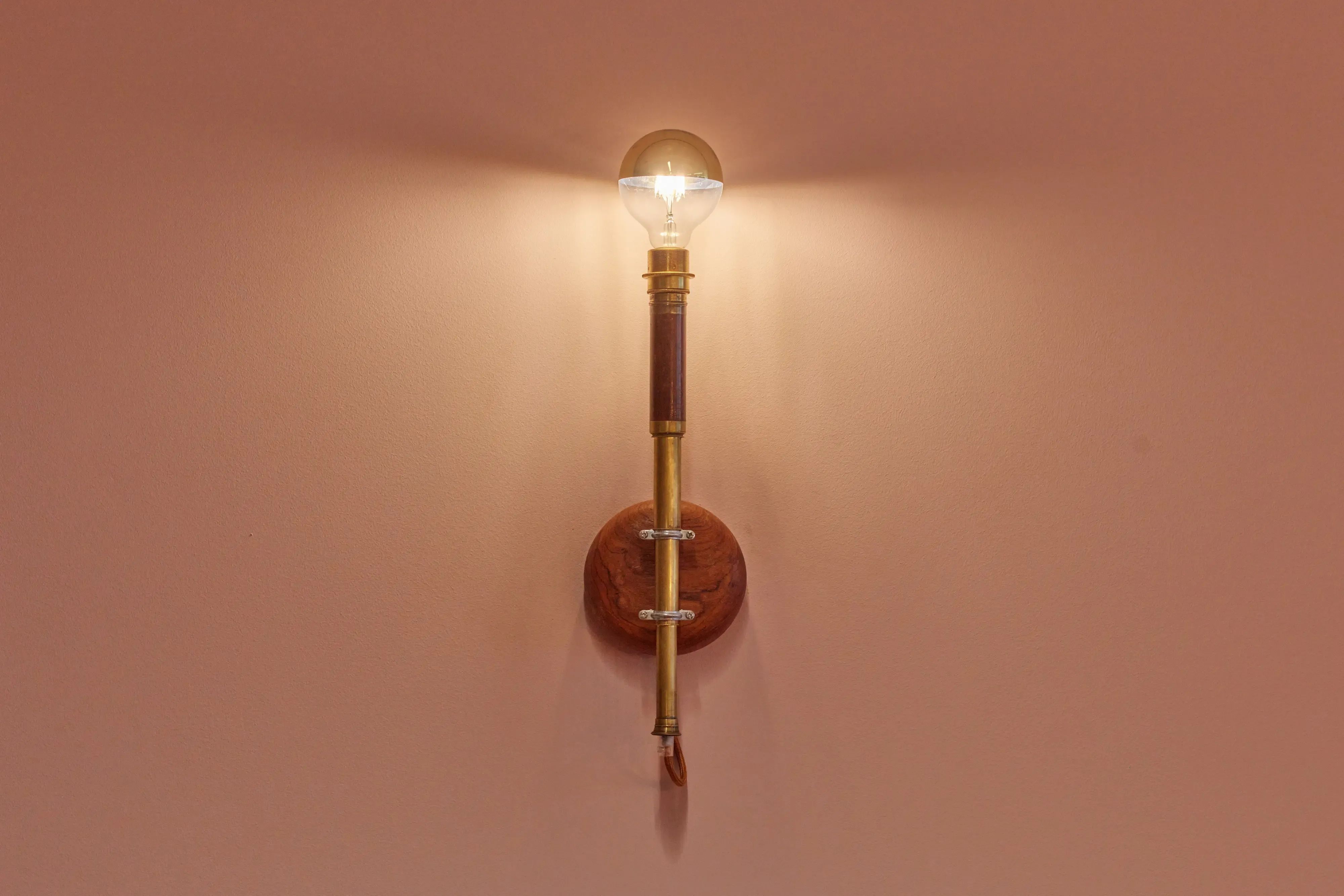 Een wandlamp met een verlichte lamp in vintage-stijl verspreidt een warme gloed. Het ontwerp van messing en hout vormt een mooie aanvulling op de zachtroze muur.
