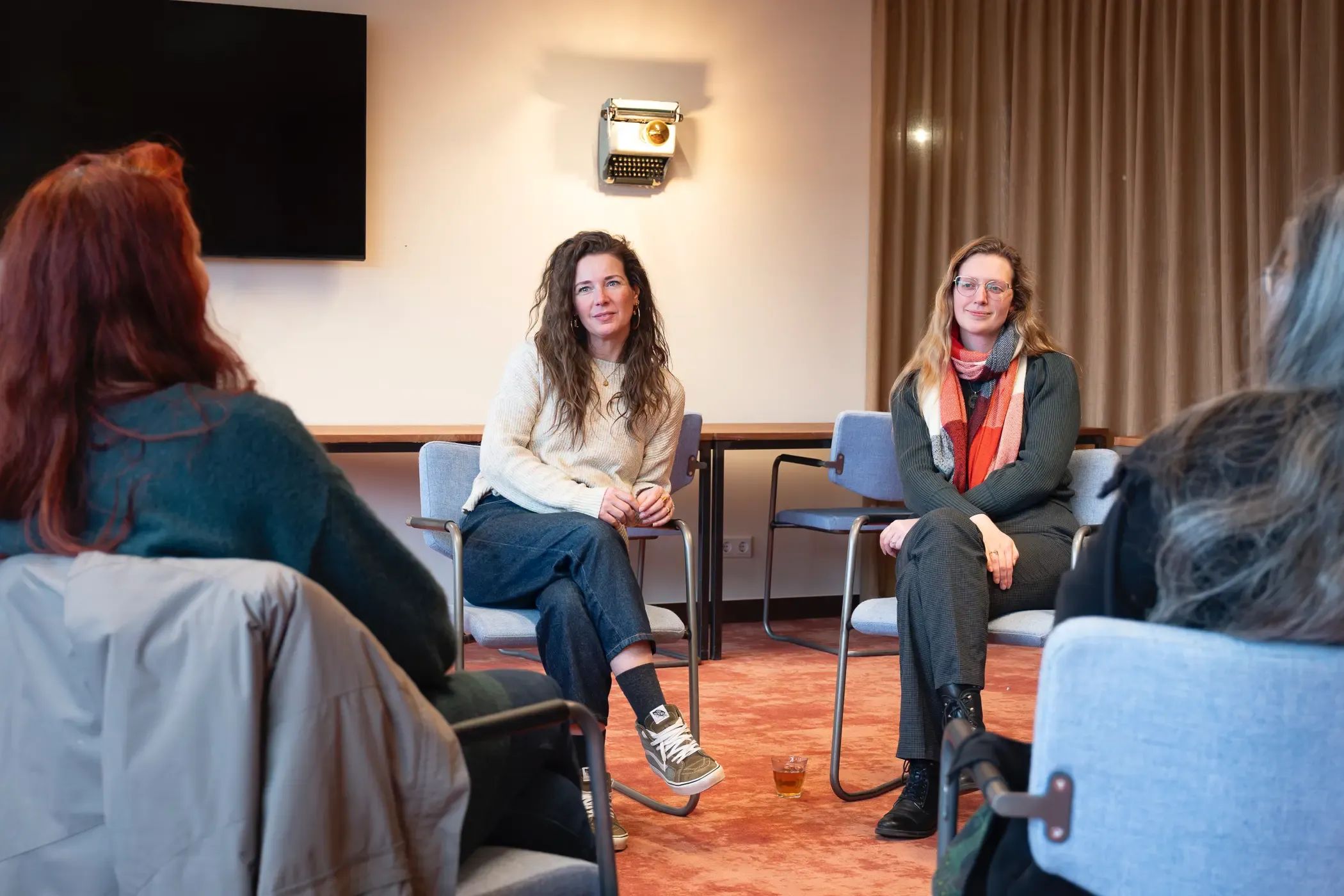 Twee vrouwen zitten tegenover een publiek en voeren een professioneel gesprek in een vergaderzaal.