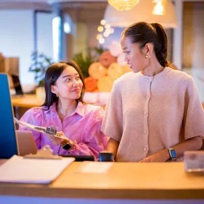 Two women collaborating at a modern office desk, one pointing at an Apple iMac screen.