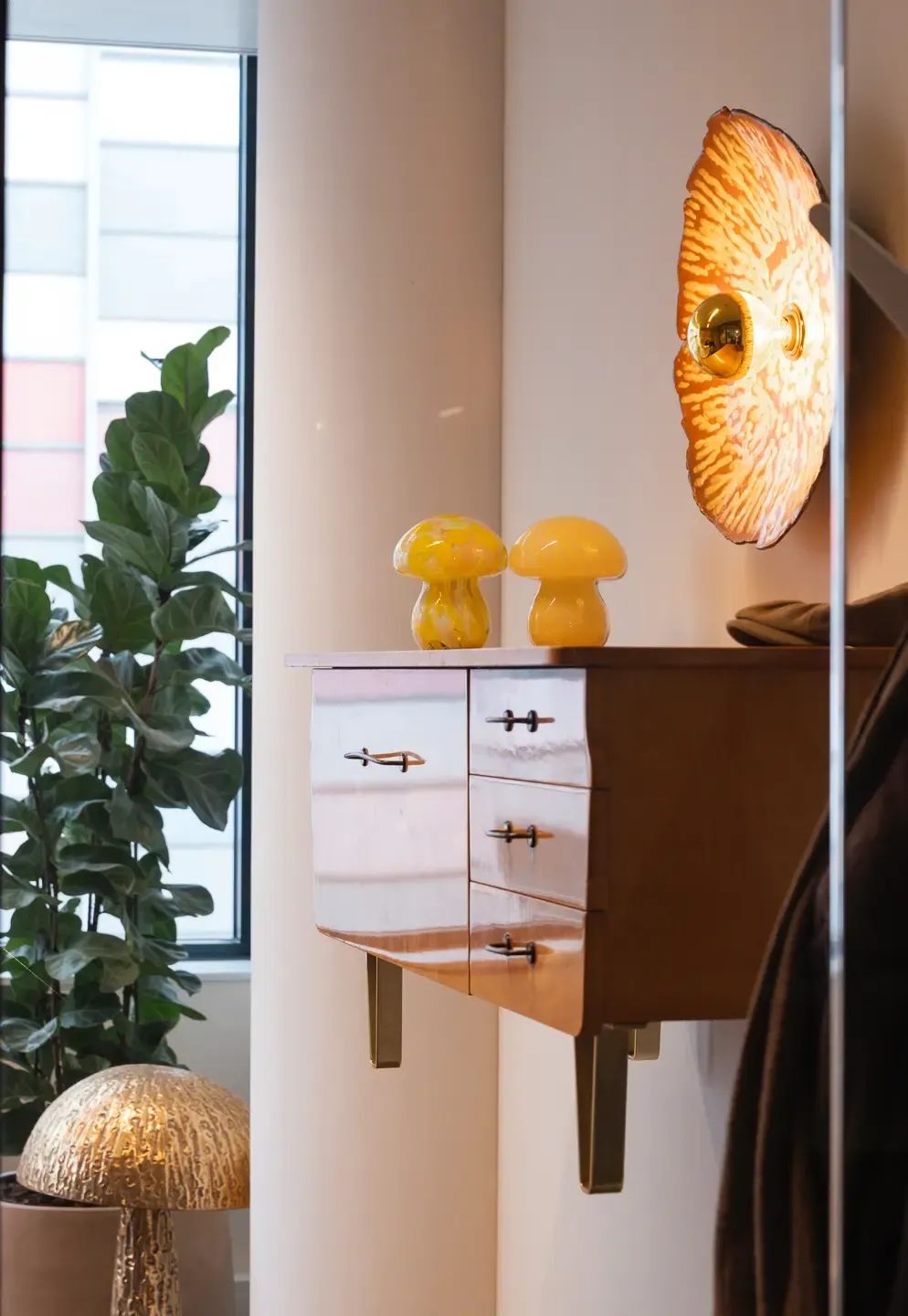 Verlichte wandlamp boven een zwevend kastje met gele paddestoelendecoratie en een groene plant.