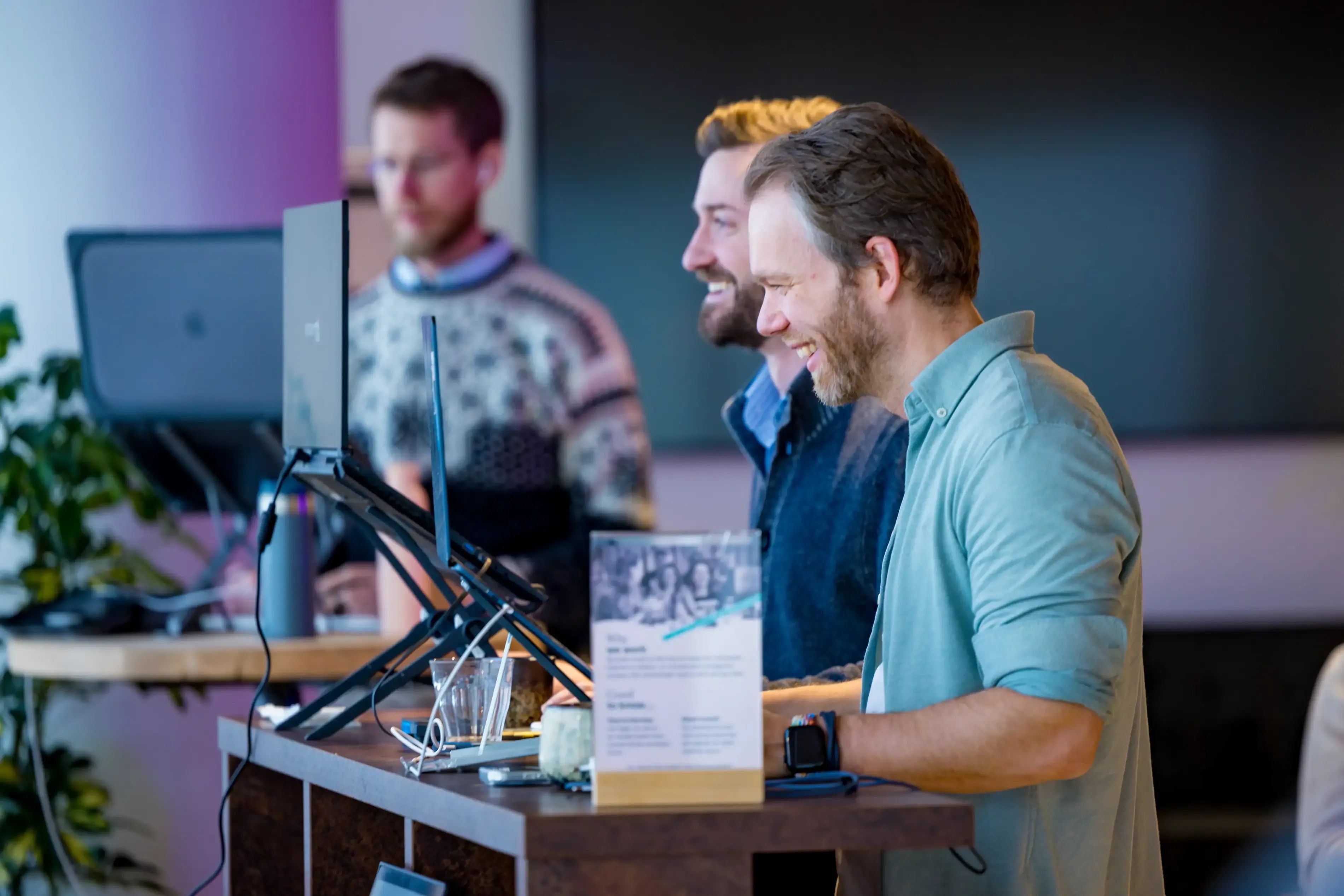 Twee glimlachende mannen bekijken tevreden iets op een toonbank, terwijl een andere man op de achtergrond aan het werk is.